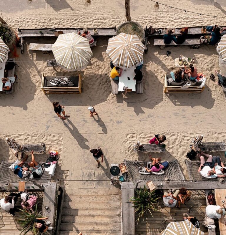 Vogelperspectief van een sfeervol strandpaviljoen met houten terrassen, gezellige zitplekken, mensen die ontspannen eten en drinken, en een verweerde boot als decoratie op het zand.